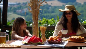 Deux personnes sont assises à une table en bois, devant de la nourriture et des boissons, à l’extérieur, avec une vue sur des collines.