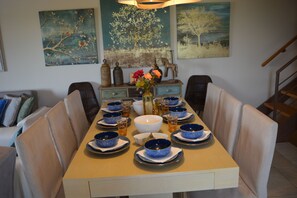 Una mesa de comedor dispuesta con cuencos azules y blancos, un florero con flores y objetos decorativos sobre una mesa de madera en un comedor con una escalera al fondo.