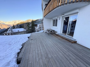 Een houten terras met een tafel en stoelen, een schuifdeur en uitzicht op besneeuwde bergen.