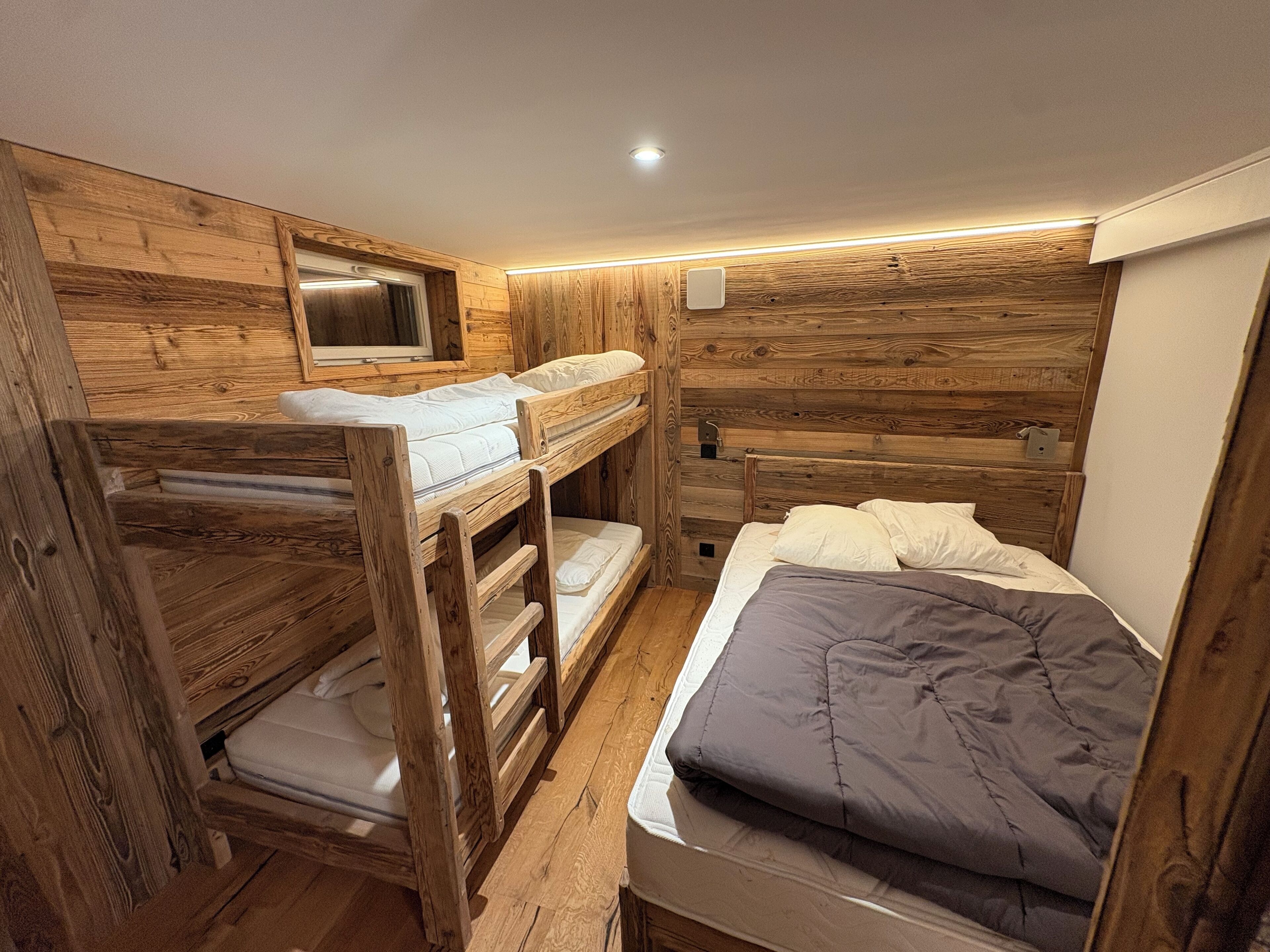 Ein Zimmer mit Holzboden, Etagenbetten, einem kleinen Fenster und einer Wandleuchte.