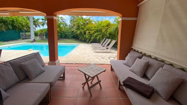 A covered outdoor seating area with two sofas and a small table, overlooking a swimming pool and palm trees.