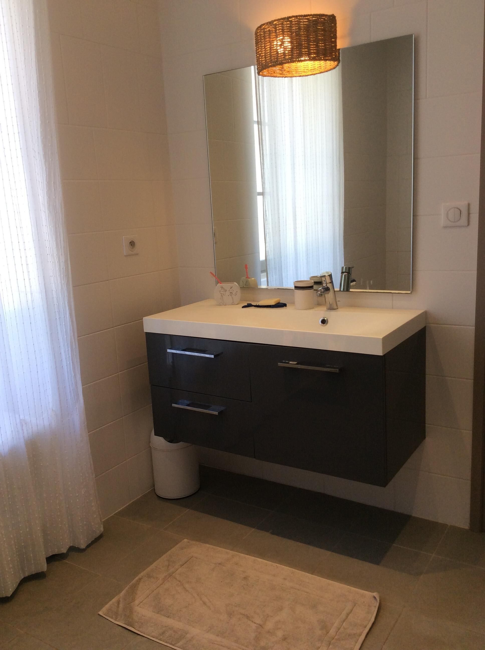 Un baño con tocador blanco, cajones oscuros, un espejo y un pequeño cubo de basura.