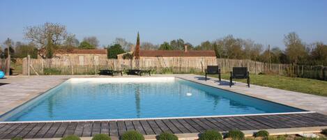 A rectangular swimming pool with a wooden deck surrounding it, a wooden fence in the background, and a clear blue sky.
