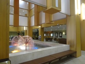 Un hall d’entrée spacieux agrémenté d’un élément décoratif central avec de l’eau, de coins salon et un comptoir d’accueil.
