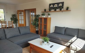 A living room with two grey sofas, a wooden coffee table, and a cabinet with glass doors. There are potted plants on the shelves and a painting of daisies on the wall.