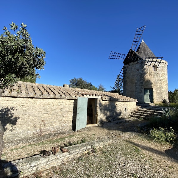 Un moulin à vent en pierre, doté d’un treuil métallique, d’une petite porte et d’un toit recouvert de tuiles.