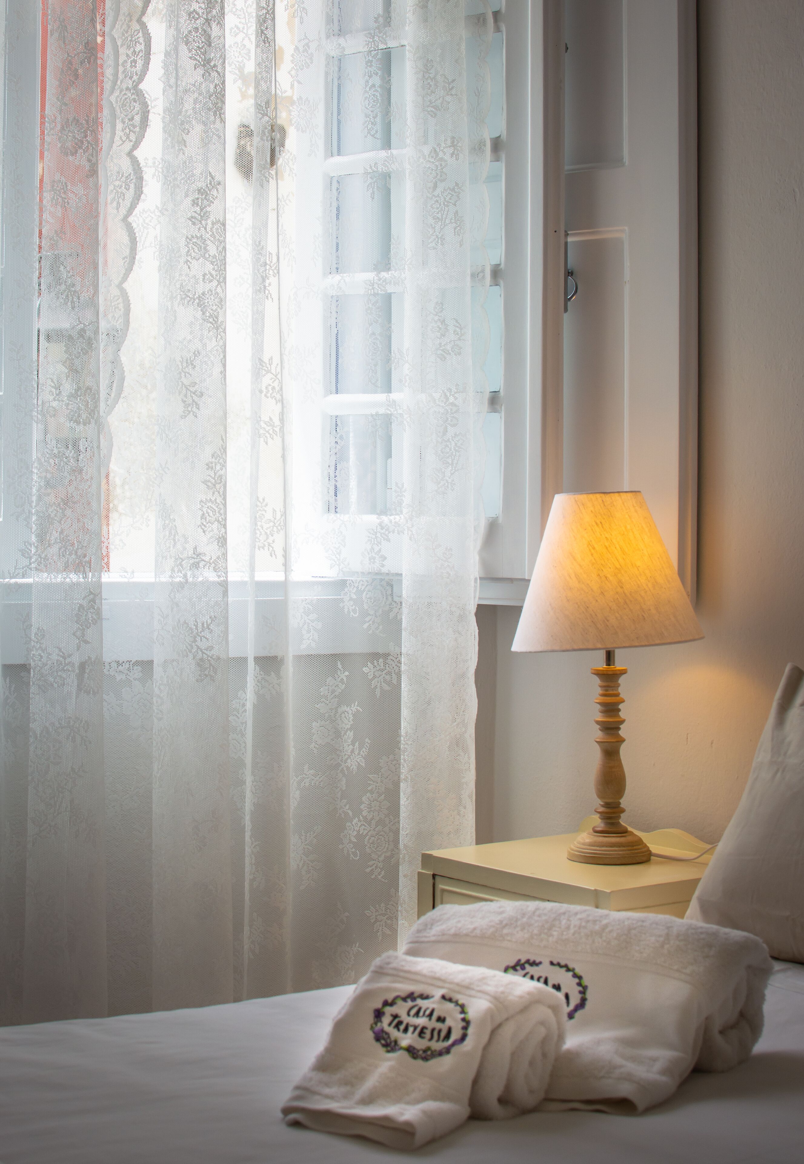 Una cama bien hecha con una toalla doblada, una lámpara de noche y cortinas traslúcidas.