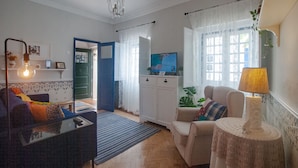 Una sala de estar con sofá, sillón, televisión y lámpara. Hay una puerta que da a otra habitación y una ventana con cortinas.
