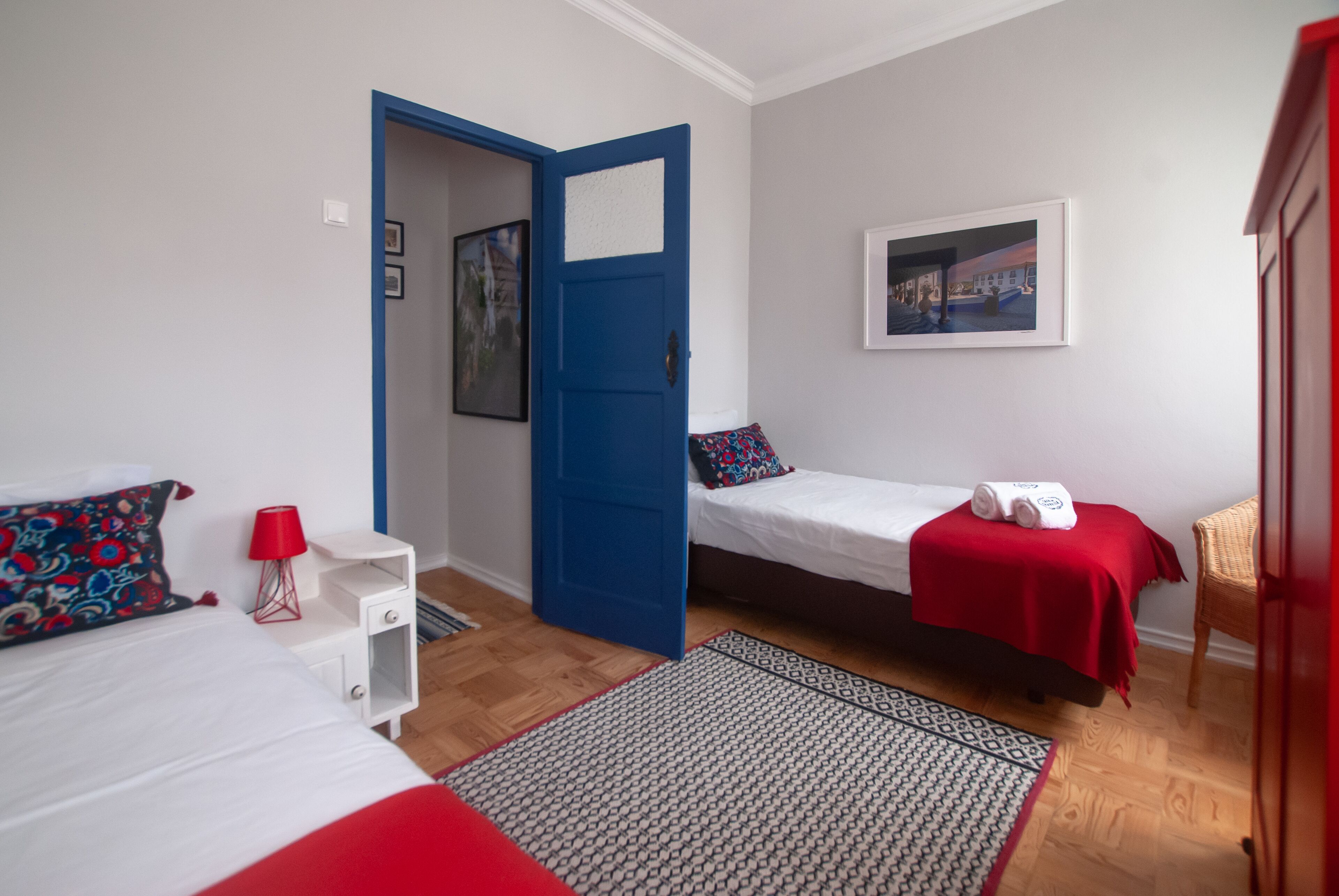 A room with two beds, a red and white bedspread, a patterned rug, a red lamp, and a blue door.