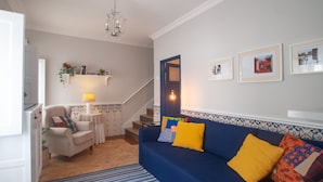 Un salón con un sofá azul, cojines amarillos, un sillón blanco, una mesita con una lámpara y una escalera que lleva a otra habitación.