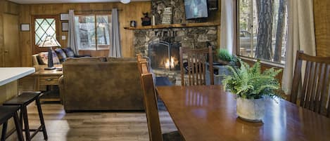 A cozy cabin interior with a stone fireplace, wooden furniture, and a large window offering a view of the forest.