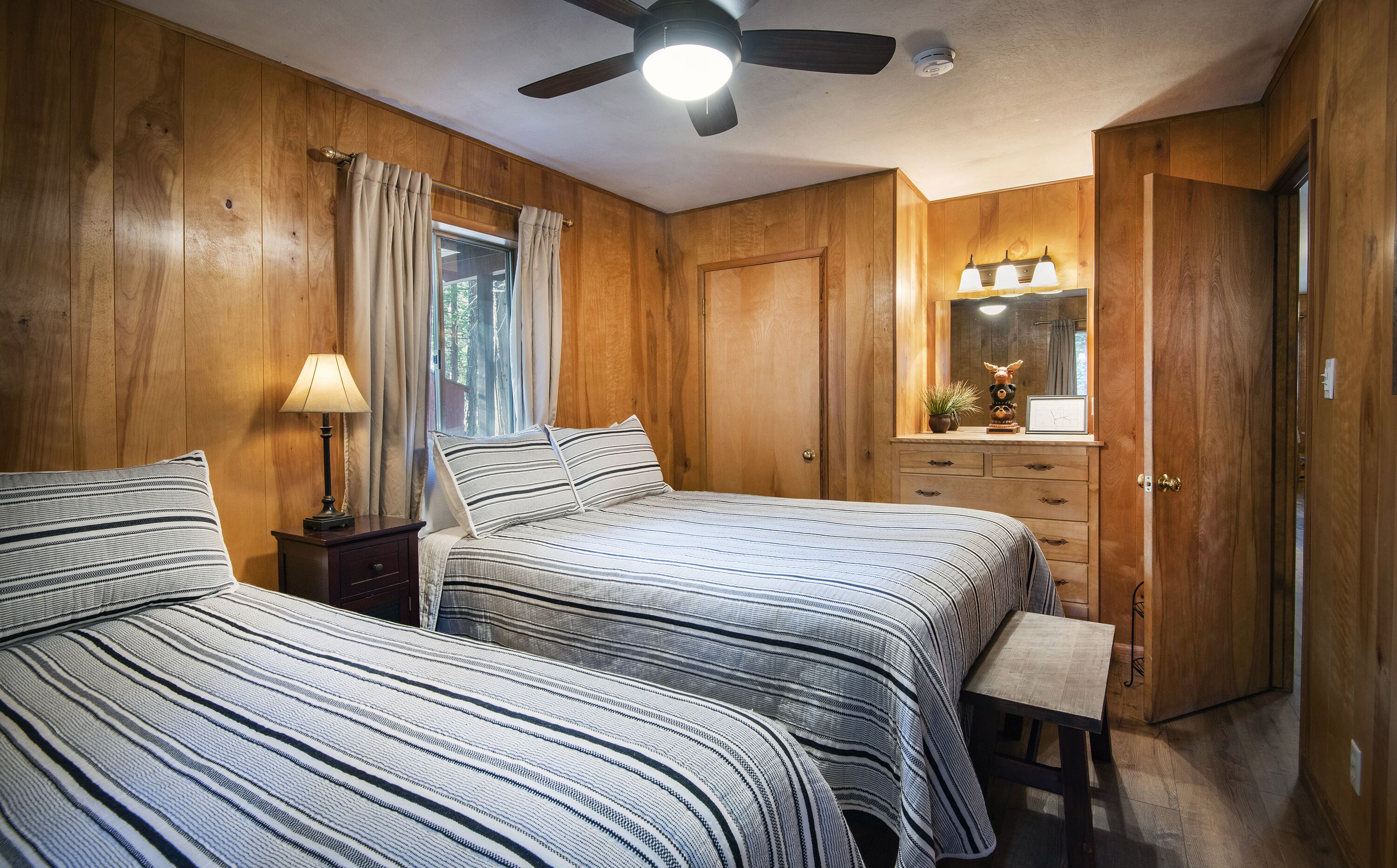 A bedroom with two beds, wooden walls, a ceiling fan, a window with curtains, a bedside table with a lamp, a dresser with drawers, a mirror, and a small stool.