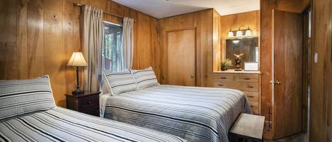 A bedroom with two beds, wooden walls, a ceiling fan, a window with curtains, a bedside table with a lamp, a dresser with drawers, a mirror, and a small stool.