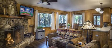 A cozy cabin interior with a stone fireplace, wooden walls, a comfortable sofa set, and a television mounted on the wall.