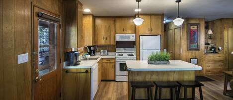 Una cocina con armarios de madera, estufa blanca, microondas, refrigerador y cafetera. Hay tres taburetes de bar y una planta en maceta en la isla.