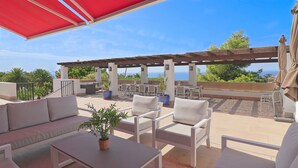 Uma área de terraço ao ar livre com móveis brancos, um toldo vermelho e vista para o mar.