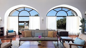 Ampla sala de estar com sofá, poltronas, mesa de centro e vista através de janelas em arco.