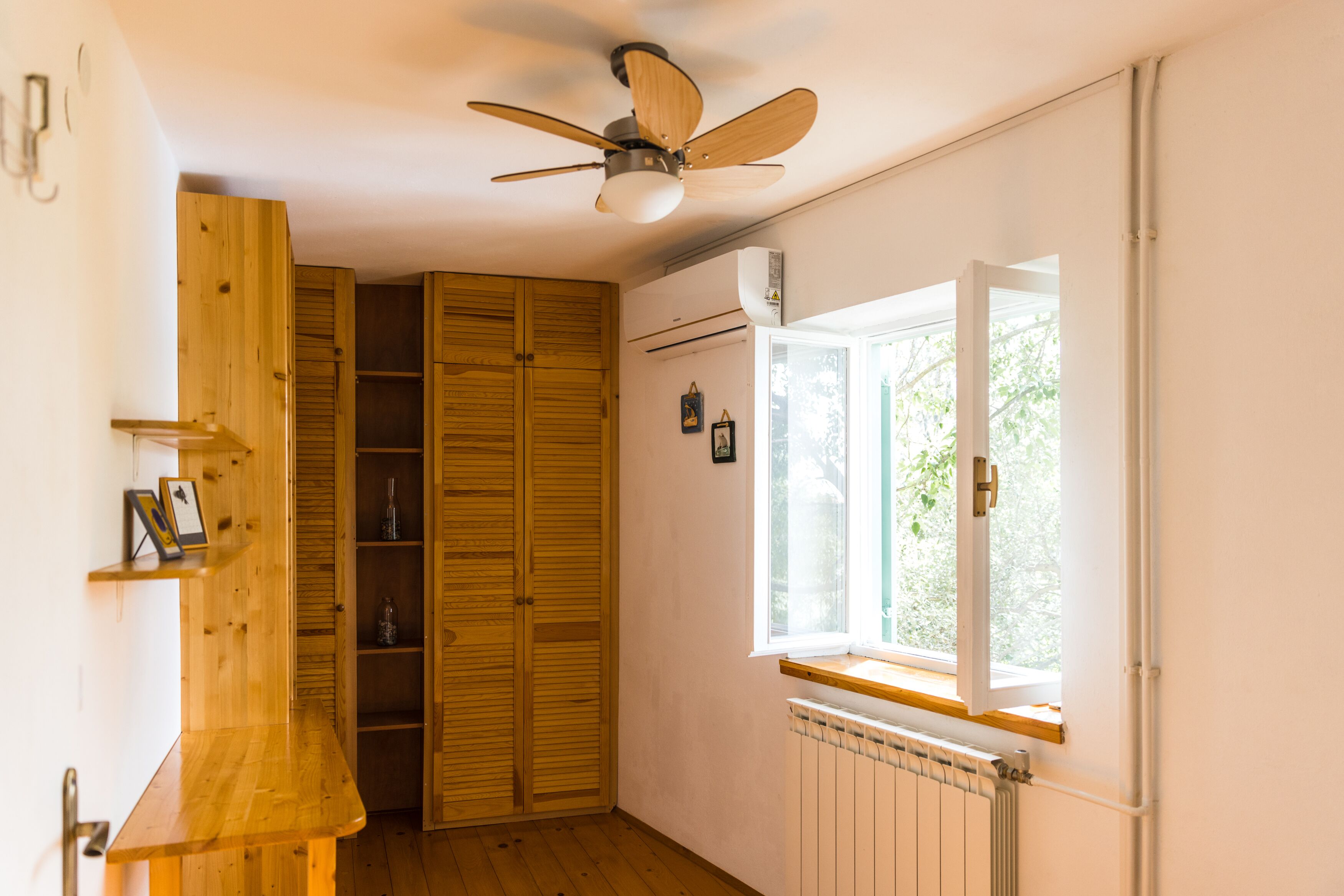 A room with a wooden wardrobe, a ceiling fan, a window with a view of greenery, and a radiator.