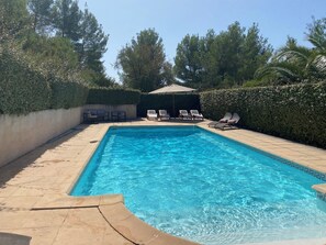 A rectangular swimming pool with clear blue water, surrounded by a tiled deck. There are lounge chairs and a small umbrella providing shade.