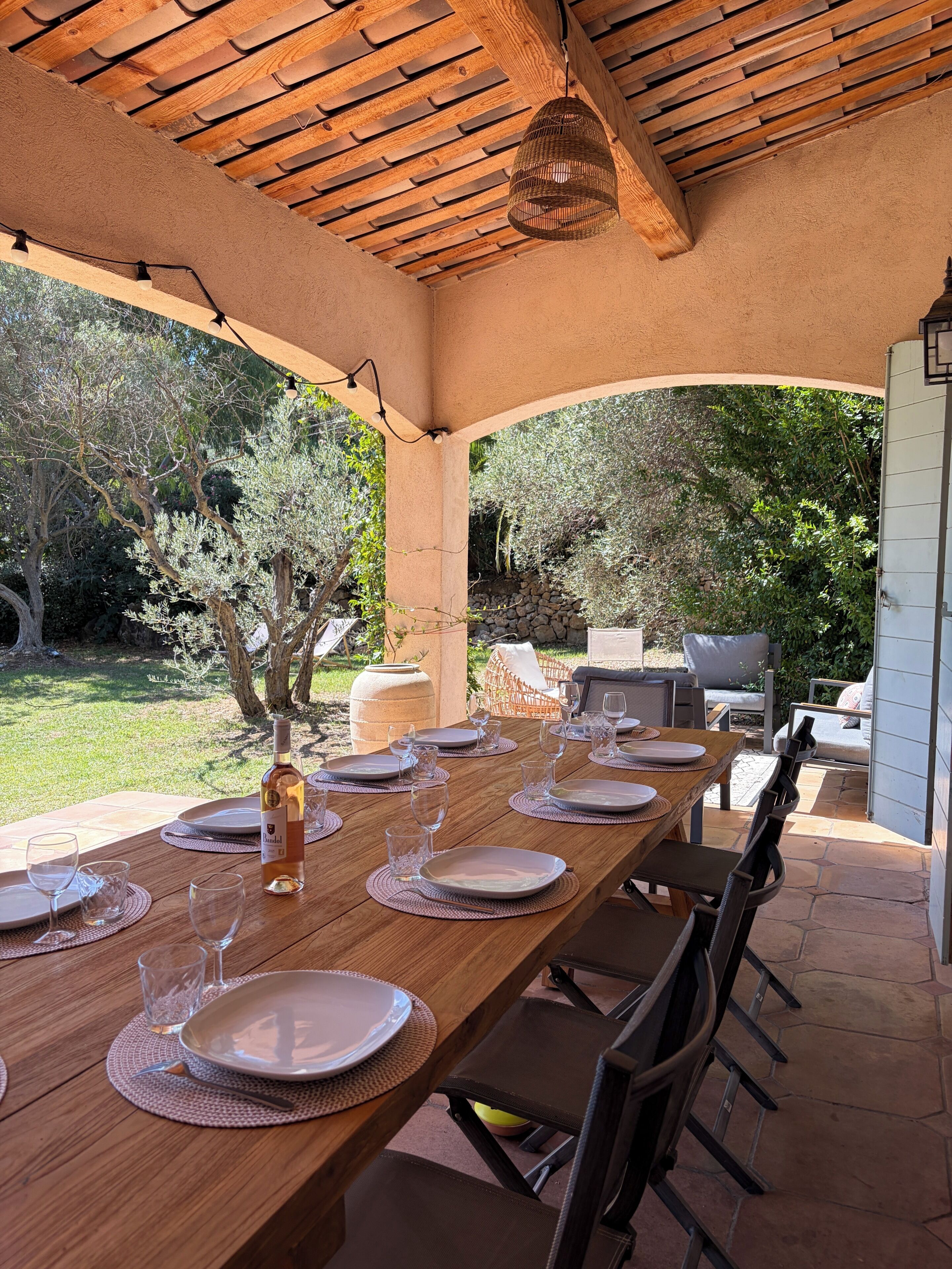 Une table à manger en bois, dressée pour six personnes avec des assiettes, des verres et une bouteille, sous un plafond en bois agrémenté d’un luminaire suspendu.