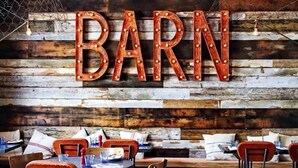A rustic interior with a wooden wall and the word "BARN" in large letters. Tables and chairs are arranged for dining. - Stunning 2 Bedroom Stone Barn with 24" Open Plan Living Room/Kitchen/Dining (Gulval)