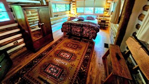 Interieur van een blokhut met een bed, een houten vloer, een plafondventilator en een raam met jaloezieën.