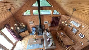 Interior acogedor de una cabaña con techo estilo altillo, paredes de madera, chimenea de piedra, un sofá, una mesa de comedor y un ventanal grande.