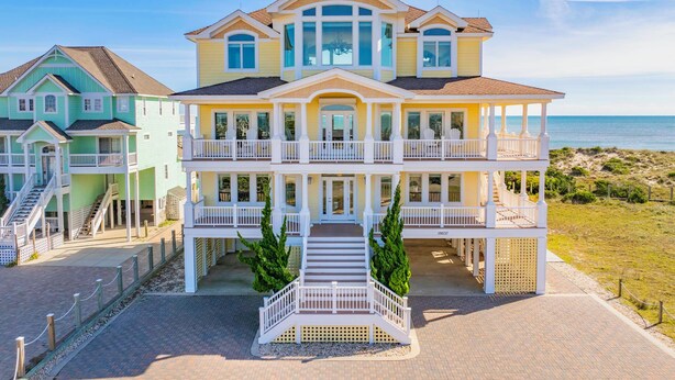 Un'ampia casa fronte spiaggia, a più piani, con un balcone centrale, un portico coperto e una scala che conduce all'ingresso.