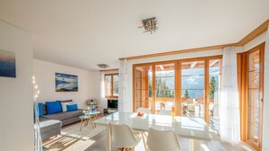 Una sala moderna con una mesa de comedor de vidrio, sillas blancas y un sofá gris con cojines azules. Hay un ventanal con marcos de madera y vistas a las montañas.