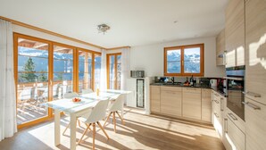 Una cocina moderna con armarios de madera, un comedor con mesa de vidrio y grandes ventanales con vistas a las montañas.