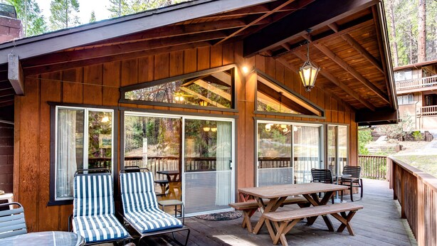 Una terraza de madera con mobiliario exterior, incluyendo sillas y mesas, y amplios ventanales que ofrecen vistas del bosque circundante.