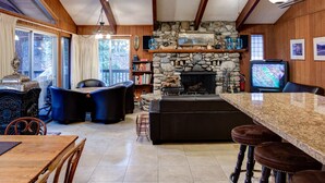 Un acogedor interior de cabaña con chimenea de piedra, vigas de madera, pared de piedra, televisor y comedor con mesa y sillas.