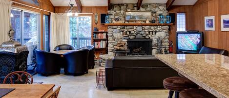 A cozy cabin interior with a stone fireplace, wooden beams, a stone wall, a television, and a dining area with a table and chairs.