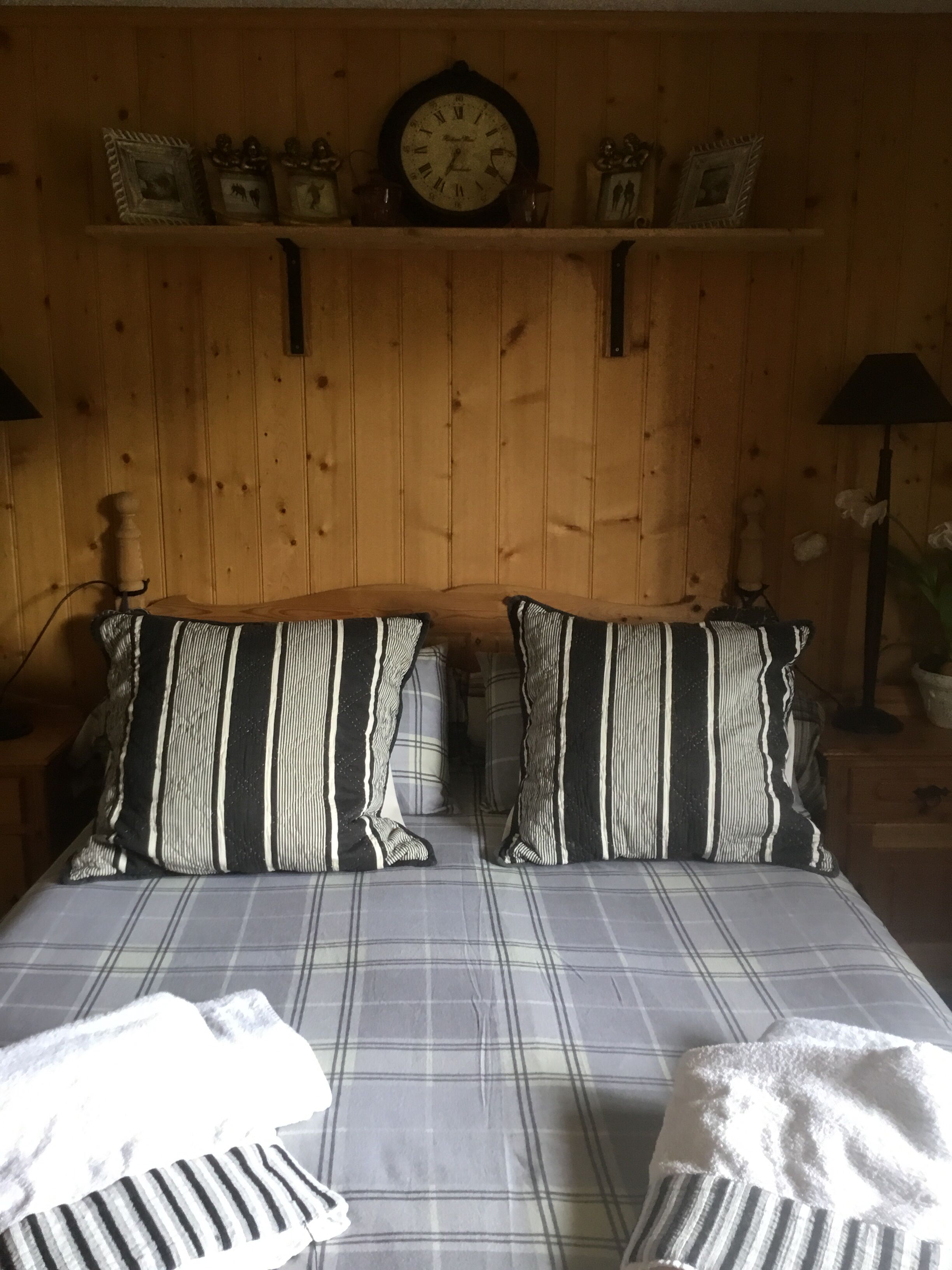 Una habitación con pared de madera, una cama con almohadas a rayas, un reloj y un estante con cuadros enmarcados.