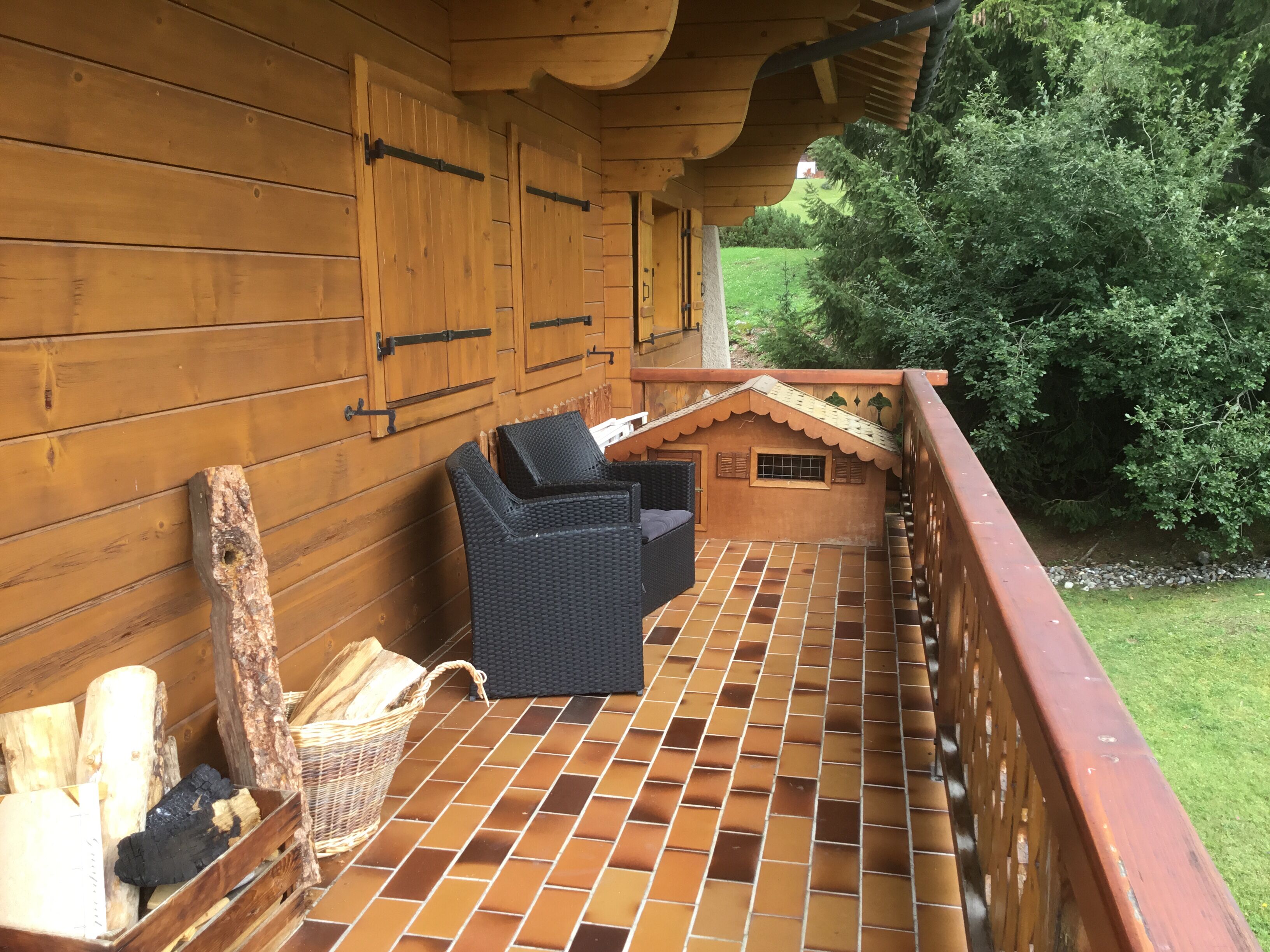 Een houten balkon met betegelde vloer, een rieten stoel en een klein houten bouwwerk met een raam.
