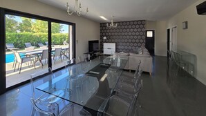 A modern living room with a glass dining table, a sofa, and a TV. There is a pool visible through the glass doors.