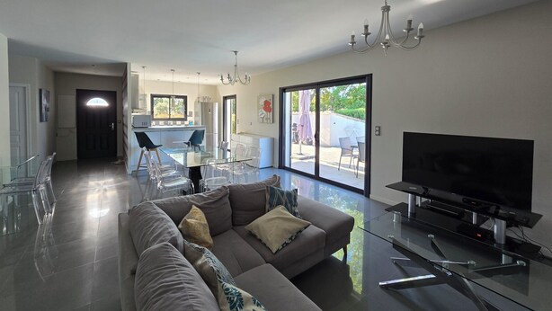 A modern living room with a glass dining table, a sofa, a flat-screen TV, and a sliding glass door leading to a patio.