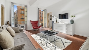 Una sala de estar moderna con sofá, una butaca roja, televisor y vistas a un paisaje urbano a través de puertas abiertas.