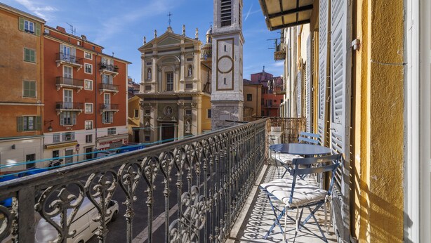 Un balcón con barandilla de hierro forjado que da a una calle con edificios y una torre del reloj.