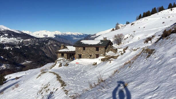 Ein Steinhaus mit schneebedecktem Dach, umgeben von schneebedeckten Hügeln und Bergen im Hintergrund.
