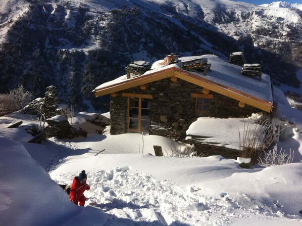 A stone cabin with a sloped roof, surrounded by snow-covered mountains.