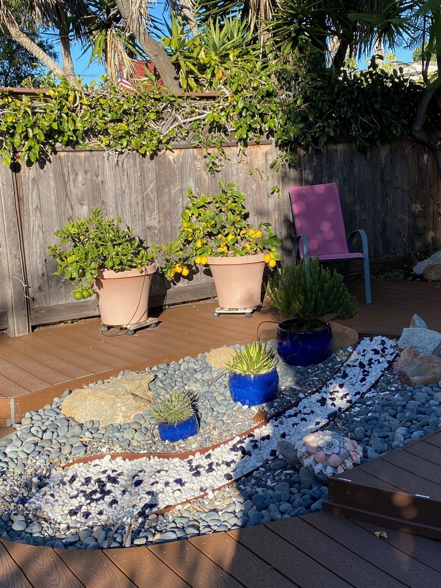 Une terrasse en bois agrémentée de plantes en pot, d’une chaise et d’un chemin décoratif fait de galets et de coquillages.