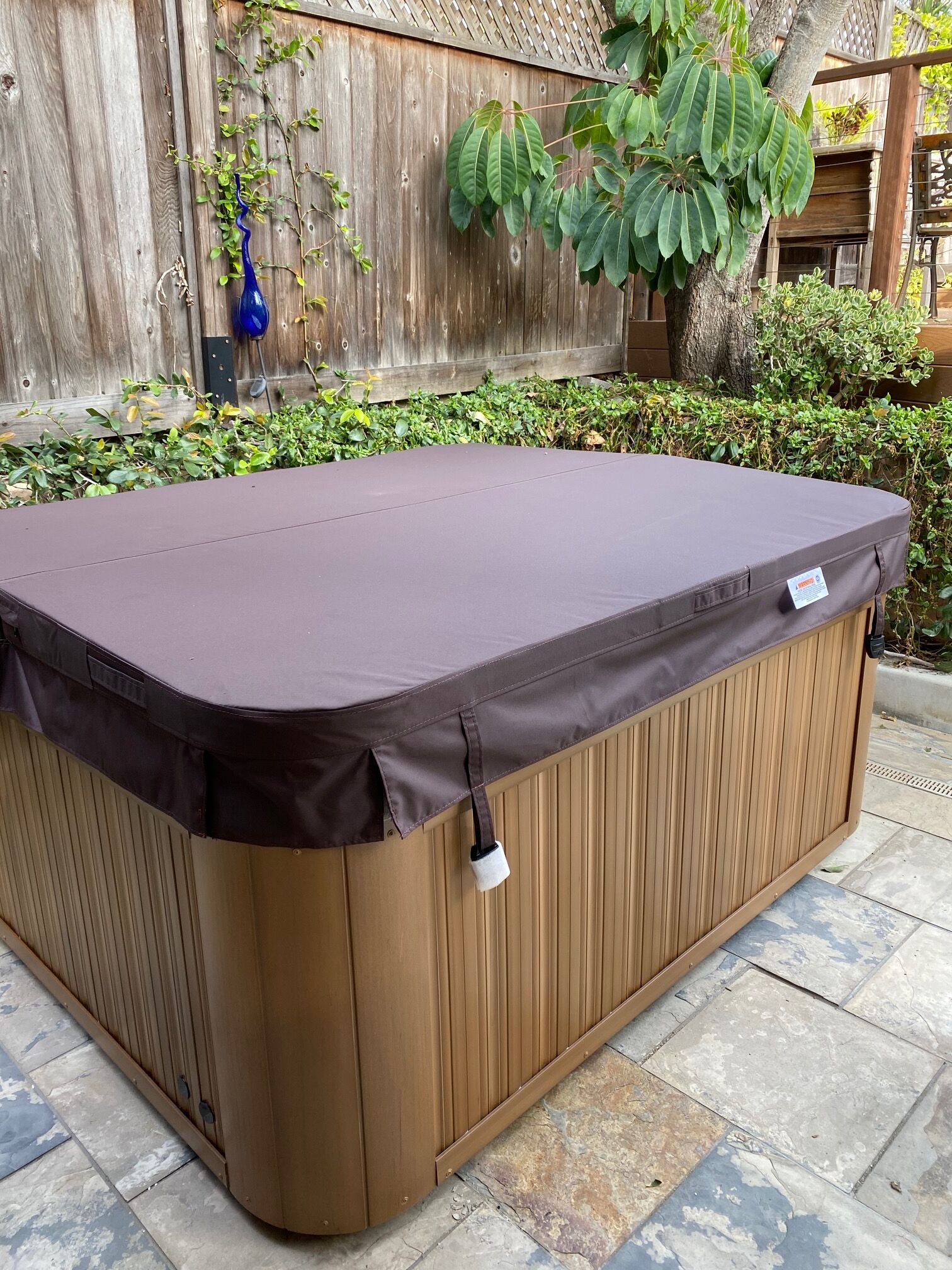 A hot tub with a brown exterior and a dark cover, placed on a tiled patio with a wooden fence and greenery in the background.