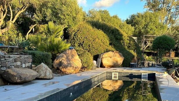 A swimming pool with a stone wall and large rocks, surrounded by lush greenery and trees. - House in natural park with river, located 15' from Tarifa and beaches <br> (TARIFA)