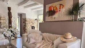 A cozy living room with a beige sofa, a fluffy throw blanket, and a straw hat on the armrest. There's a vase with flowers on a wooden table, and a stone wall in the background. - House in natural park with river, located 15' from Tarifa and beaches <br> (TARIFA)
