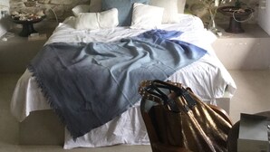A bedroom with a stone wall, a bed with white and blue bedding, a wooden chair, and a bedside table with a lamp. - House in natural park with river, located 15' from Tarifa and beaches <br> (TARIFA)