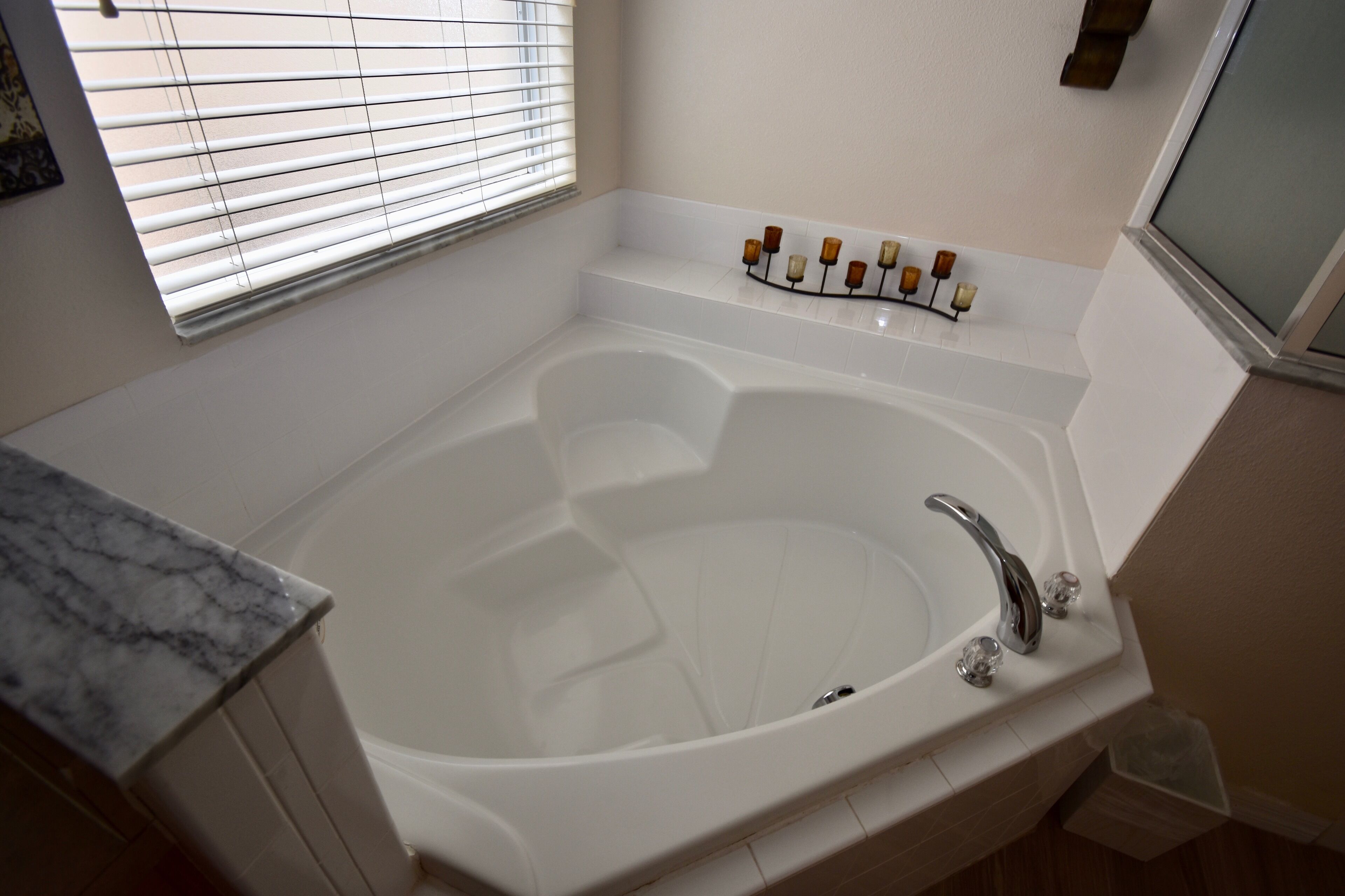 A bathroom with a large bathtub, a marble countertop, and a window with blinds.