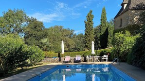 Una piscina rectangular con agua azul cristalina, rodeada de sillas de descanso y sombrillas, con árboles y un muro de piedra al fondo.