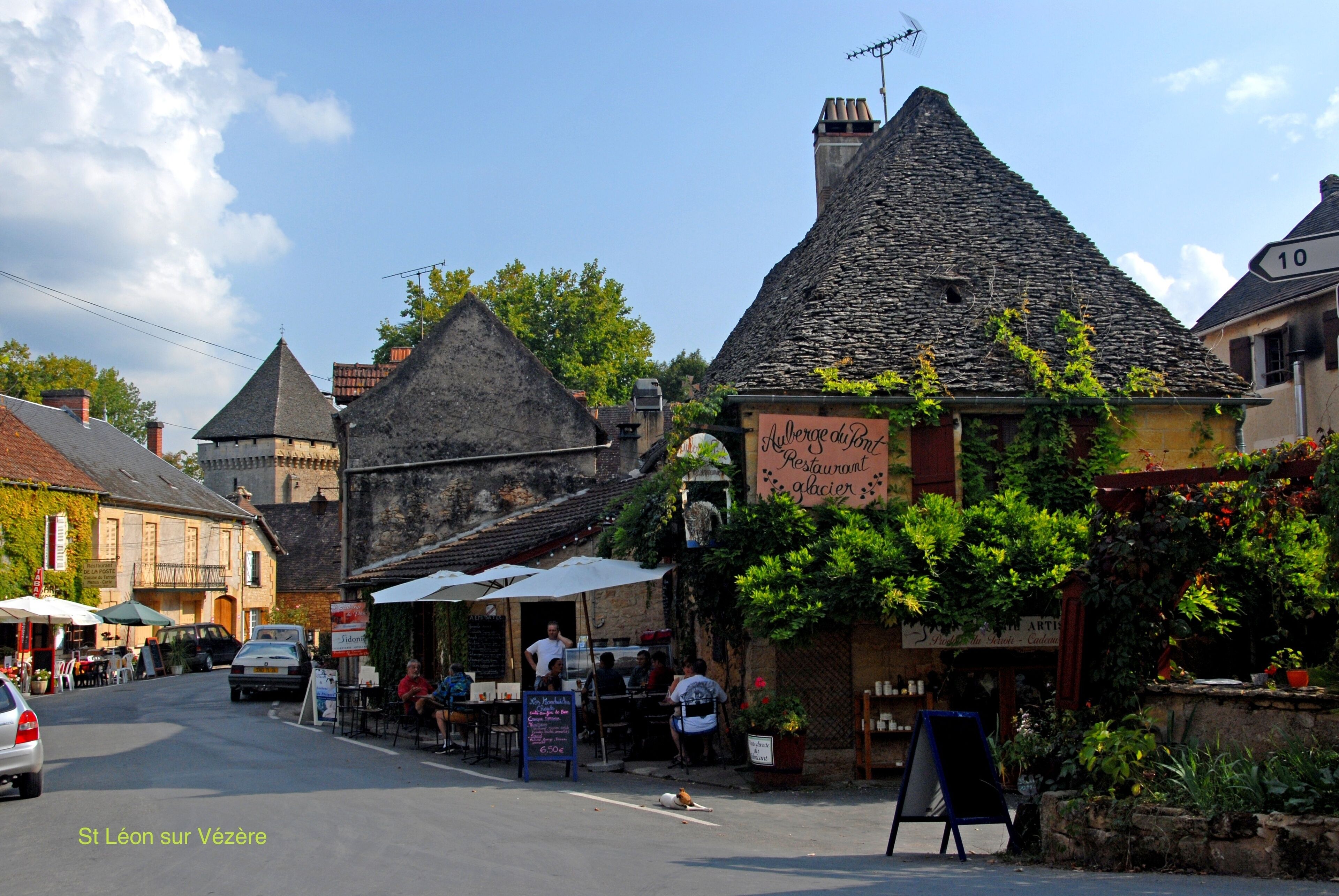 Une scène de rue à Saint-Léon-sur-Vézère, avec un restaurant appelé « Auberge du Pont Restaurant Glacier » et des tables en terrasse.