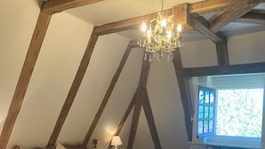 Un dormitorio con techo inclinado, vigas de madera a la vista, una araña, una cama con ropa de cama blanca, una mesita de noche y una estantería.